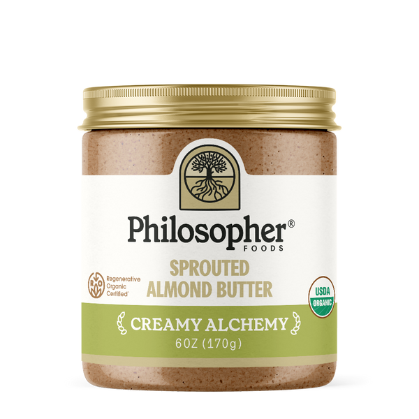 Image displays front of jar. Label displays image of tree with roots, "Philosopher Foods" in bold black text, "Sprouted Almond Butter" in gold, "Creamy Alchemy" in white, 6 ounce jar. Also shows label icons for USDA Certified organic and for Regenerative Organic Certified
